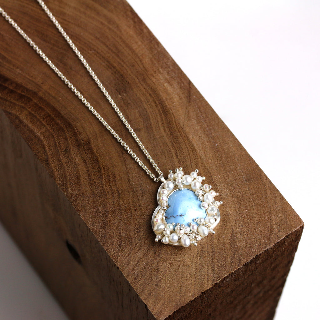 Turquoise heart pendant surrounded by clustered pearls on a silver chain on wood block.