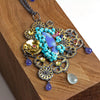 An ornate pendant necklace displayed on a wooden block. Design features opal, turquoise, gold and blackened silver.